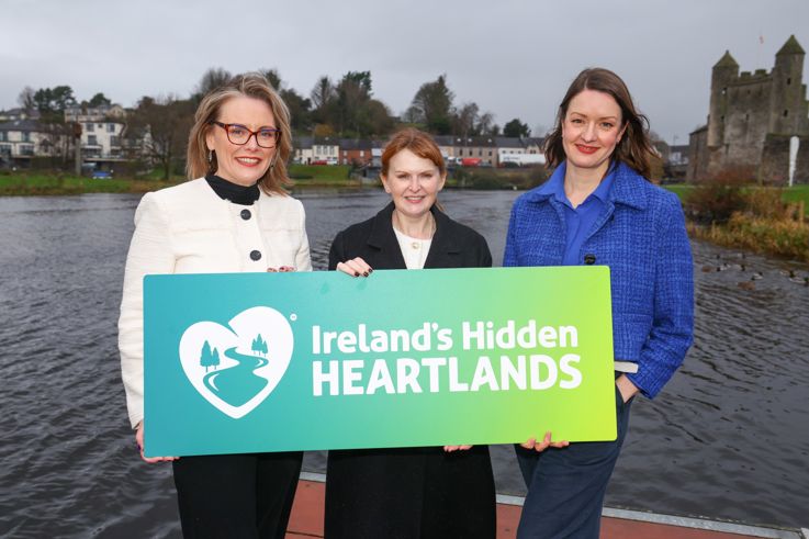  Laura McCorry, Chief Executive of Tourism Northern Ireland, Economy Minister Caoimhe Archibald and Alice Mansergh, Chief Executive of Tourism Ireland standing at a picturesque landscape representing Ireland's Hidden Heartlands, featuring green fields and serene lakes.
